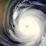 Immagine fotorealistica del Golfo del Bengala visto dall'alto: un'area con acqua superficiale visibilmente più calda (leggera distorsione termica o colore leggermente diverso) accanto a un ciclone tropicale in formazione, nuvole vorticose intense ma localizzate. Obiettivo zoom teleobiettivo 150mm, ripresa aerea, alta definizione, luce drammatica del tardo pomeriggio.