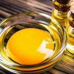 Primo piano fotorealistico di un tuorlo d'uovo diviso che mostra la sua ricca consistenza dorata, accanto a piccole fiale di vetro trasparente contenenti olio di lino e olio di girasole su una superficie di legno scuro. Obiettivo macro, 80mm, alta definizione, illuminazione laterale controllata che enfatizza la ricchezza nutrizionale e la lucentezza degli oli.