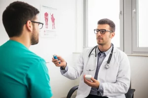 Fotografia realistica in stile reportage, un medico di base nel suo studio nel Sud Italia discute con un paziente asmatico, mostrando un inalatore. Sullo sfondo, poster medici e una finestra. Obiettivo 50mm, luce ambientale, profondità di campo media.