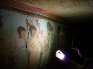 Scatto grandangolare, obiettivo 24mm, all'interno di un'antica grotta cinese scarsamente illuminata (simile a Mogao). Il focus è su una sezione di un vibrante murale che mostra segni di fessurazioni dovute all'invecchiamento. Una mano di un ricercatore (fuori fuoco) tiene una lampada UV che illumina una piccola area, rivelando sottili linee fluorescenti lungo le crepe. Profondità di campo, illuminazione drammatica.