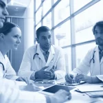 Un gruppo di giovani medici specializzandi in pediatria, con camici bianchi, che discutono animatamente attorno a un tavolo rotondo su cui sono appoggiate pubblicazioni mediche e un tablet, in un ambiente ospedaliero moderno e luminoso. Obiettivo zoom 24-70mm impostato a circa 35mm, luce naturale che entra da una grande finestra laterale, espressioni concentrate ma collaborative, profondità di campo media per mantenere a fuoco il gruppo e sfocare leggermente lo sfondo dell'ospedale. Duotone blu e grigio.