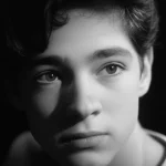 Ritratto fotografico di un adolescente con espressione pensierosa e malinconica, luce laterale che crea ombre profonde sul viso, obiettivo 50mm, stile film noir, bianco e nero con leggera grana, profondità di campo ridotta per sfocare lo sfondo e concentrare l'attenzione sul soggetto.