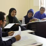 Un gruppo diversificato di studenti universitari della Edna Adan University in Somaliland, alcuni impegnati in una discussione animata e altri che studiano individualmente in un'aula luminosa. Portrait photography, prime lens 35mm, depth of field, luce naturale che entra dalle finestre, per trasmettere un senso di ambiente accademico e le diverse esperienze degli studenti.