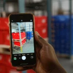 Primo piano di una mano che tiene uno smartphone con un'app di logistica commerciale aperta, sullo sfondo sfocato si intravedono casse di bevande analcoliche pronte per l'esportazione in un magazzino dell'Africa Occidentale. Obiettivo 50mm, profondità di campo, illuminazione industriale calda.