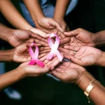 Un'immagine simbolica che rappresenta l'equità nelle cure mediche per il cancro al seno metastatico: mani di diverse etnie che si sostengono a vicenda, con un nastro rosa stilizzato sullo sfondo. Prime lens, 35mm, depth of field, colori caldi e rassicuranti.