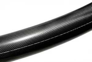 Vista leggermente angolata di un segmento di pannello cilindrico in materiale composito avanzato (fibra di carbonio) con un intaglio di forma ellittica decentrato. L'illuminazione laterale evidenzia la curvatura e la trama del materiale. Obiettivo prime 35mm, profondità di campo ridotta che sfoca leggermente lo sfondo, stile fotorealistico industriale.