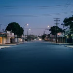 Immagine simbolica dell'impatto della deindustrializzazione e del COVID-19 su una comunità australiana: una strada deserta in una cittadina industriale al crepuscolo, con le luci delle case accese ma poche persone visibili. Wide-angle lens, 20mm, long exposure, atmosfera malinconica ma con un barlume di speranza, colori freddi con accenti caldi dalle finestre.