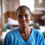 A photorealistic image, 35mm portrait lens, showing a compassionate midwife in a Ugandan hospital maternity ward, looking directly at the viewer. The background is slightly blurred (depth of field) to focus on her, hinting at the busy environment she works in. The lighting is soft but realistic.