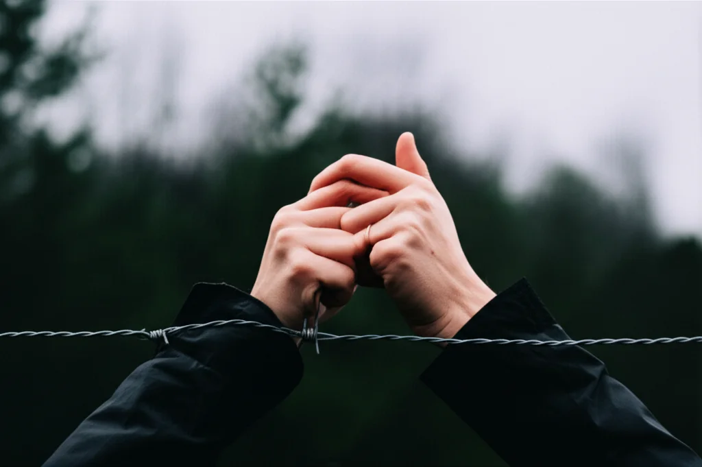 Immagine fotorealistica e concettuale delle mani che trema su una recinzione a filo spinato, simboleggiano dialoghi difficili e cercano una connessione in ambienti ostili, illuminazione drammatica, obiettivo da 50 mm, apertura f/3.5