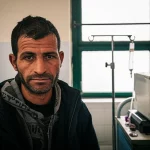 Immagine emblematica di un paziente siriano durante una seduta di emodialisi in un centro medico improvvisato nel Nord-Ovest della Siria. Il paziente guarda fuori da una finestra, espressione mista di stanchezza e speranza. Accanto, la macchina per la dialisi è in funzione. L'ambiente mostra segni del conflitto (muri scrostati, finestre protette) ma è pulito. Luce naturale filtrata, obiettivo 35mm, profondità di campo che isola il paziente ma lascia intravedere il contesto. Photorealistic, portrait photography, 35mm prime lens, depth of field, natural light.