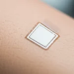 Fotografia macro di un sensore indossabile flessibile basato su nanotubi di nitruro di boro (BNNT/PDMS), applicato delicatamente sulla pelle vicino a un'articolazione del ginocchio umano in leggero movimento. Obiettivo macro 90mm, illuminazione controllata e diffusa per evidenziare i dettagli fini del materiale e la sua perfetta conformabilità alla curvatura della pelle, messa a fuoco estremamente precisa sul sensore, sfondo neutro e sfocato.
