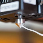 Fotografia macro di un filamento bianco opaco di PHB/amido che esce dall'estrusore caldo di una stampante 3D FDM durante il processo di stampa. Dettagli nitidi sul filamento fuso e sulla punta dell'ugello metallico. Obiettivo macro 105mm, illuminazione controllata per evidenziare la texture plastica, sfondo leggermente sfocato della macchina in funzione.