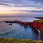 Veduta aerea delle Isole Magdalen, Canada, al tramonto. Le scogliere rosse di arenaria contrastano con il blu intenso del Golfo di San Lorenzo. Obiettivo grandangolare 14mm, lunga esposizione per enfatizzare il movimento delle nuvole e la levigatezza dell'acqua, luce dorata, alta definizione, che suggerisce la vulnerabilità delle risorse idriche costiere.