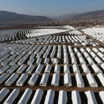 Fotografia aerea di un vasto campo di tende bianche per sfollati allestito in una piana vicino a una città parzialmente distrutta dopo un terremoto, provincia di Hatay. Obiettivo grandangolare 10-24mm, luce del tardo pomeriggio che crea lunghe ombre, messa a fuoco nitida su tutto il campo.