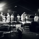 Fotografia realistica di un'unità di terapia intensiva affollata e con risorse limitate in una zona di guerra, medici e infermieri al lavoro con attrezzature essenziali, luce fioca, obiettivo prime 35mm, bianco e nero con forte contrasto, stile reportage.
