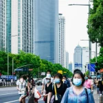 Fotografia di strada di una via di Shenzhen con persone che indossano mascherine durante la pandemia, alcuni grattacieli moderni sullo sfondo. Obiettivo 35mm, colori vivaci ma con un'atmosfera di cautela, profondità di campo.