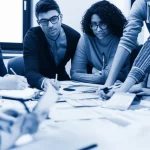 Immagine fotorealistica, obiettivo prime 35mm, stile duotone blu e grigio. Un gruppo misto di studenti e docenti del settore sanitario lavora attivamente insieme a un tavolo pieno di schemi e appunti, co-progettando materiale didattico. Profondità di campo che mette a fuoco l'interazione collaborativa e l'atmosfera di partnership.