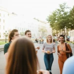 Un gruppo eterogeneo di giovani adulti Millennial che interagiscono amichevolmente in un quartiere urbano moderno e vivace, con architettura mista e spazi verdi. Luce naturale del tardo pomeriggio, effetto profondità di campo. Obiettivo prime 35mm.