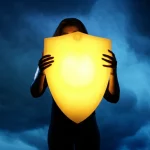 Immagine concettuale di una persona che affronta una tempesta emotiva con uno scudo luminoso che rappresenta la tolleranza allo stress. Stile fotorealistico, obiettivo prime 35mm, effetto profondità di campo, toni bicromatici freddi (blu) e caldi (giallo) a contrasto per simboleggiare la lotta e la resilienza.