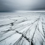 Wide-angle landscape, 15mm, vista espansiva di una banchisa di ghiaccio marino artico con strati e texture visibili, alcune aree mostrano macchie più scure indicative di sedimenti o contaminanti intrappolati, sotto un cielo freddo e drammatico, lunga esposizione per acqua liscia attorno alla banchisa, messa a fuoco nitida sui dettagli del ghiaccio.
