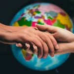 Primo piano di una mano che ne stringe un'altra con delicatezza, sullo sfondo sfocato un globo terrestre illuminato in modo non uniforme con aree più scure e più chiare a simboleggiare la distribuzione diseguale della sofferenza, fotografia macro, 85mm, illuminazione controllata laterale per creare contrasto, alta definizione, toni caldi e umani.