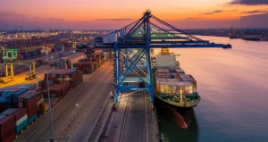 Veduta aerea di un moderno porto container dell'Africa Occidentale al tramonto, gru illuminate che caricano/scaricano una grande nave portacontainer, pile ordinate di container sullo sfondo. Obiettivo grandangolare 10-24mm, lunga esposizione per acqua liscia, colori caldi del tramonto, focus nitido.