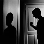 Immagine simbolica della violenza domestica da figlio a genitore: una porta socchiusa da cui si intravede un'ombra tesa tra un giovane e un adulto in un interno svedese. Film noir, prime lens 35mm, forte contrasto, bianco e nero.