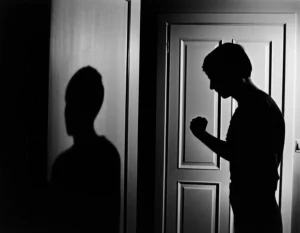 Immagine simbolica della violenza domestica da figlio a genitore: una porta socchiusa da cui si intravede un'ombra tesa tra un giovane e un adulto in un interno svedese. Film noir, prime lens 35mm, forte contrasto, bianco e nero.