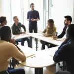 Fotografia realistica di un'aula universitaria moderna e luminosa, studenti diversi e attivamente impegnati in discussioni di gruppo animate attorno a tavoli rotondi, con un professore che facilita la conversazione spostandosi tra i gruppi. Obiettivo prime, 35mm, profondità di campo che mette a fuoco un gruppo in primo piano lasciando gli altri leggermente sfocati, luce naturale abbondante da ampie finestre, atmosfera collaborativa ed energica.