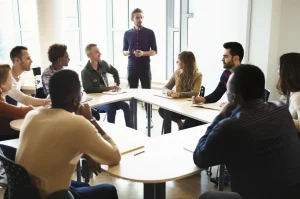Fotografia realistica di un'aula universitaria moderna e luminosa, studenti diversi e attivamente impegnati in discussioni di gruppo animate attorno a tavoli rotondi, con un professore che facilita la conversazione spostandosi tra i gruppi. Obiettivo prime, 35mm, profondità di campo che mette a fuoco un gruppo in primo piano lasciando gli altri leggermente sfocati, luce naturale abbondante da ampie finestre, atmosfera collaborativa ed energica.