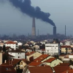 Immagine fotorealistica di un'area residenziale vicino a un sito industriale da cui si leva ancora un filo di fumo scuro, simbolo dell'impatto psicologico persistente di un incendio. Obiettivo grandangolare, 10-24mm, per catturare l'ampiezza della scena e il contrasto tra case e industria, messa a fuoco nitida.