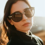 Una persona indossa occhiali da sole protettivi avvolgenti e scuri, con il sole che splende leggermente alle sue spalle, creando un leggero effetto controluce. Il focus è sugli occhiali e sulla loro aderenza al viso. Prime lens, 35mm, depth of field, luce naturale e calda.