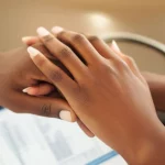 Primo piano di due mani che si stringono delicatamente, una maschile e una femminile, di etnia africana, simboleggiando l'unione e la cura, con sullo sfondo sfocato un certificato medico o un referto di screening genetico. Macro lens, 85mm, high detail, luce soffusa e calda che illumina le mani, evocando speranza e responsabilità condivisa in un contesto di pianificazione familiare.