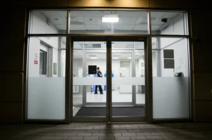 Photorealistic wide-angle 24mm shot of an emergency department entrance at night, showing the automatic doors and a few staff members visible inside, conveying a sense of constant readiness and the environment where violence can occur. Controlled lighting, sharp focus.