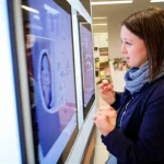 Un collage di immagini che mostra diverse persone interagire con display pubblici intelligenti in un ambiente universitario. Alcuni usano una carta RFID, altri guardano uno schermo con un'interfaccia di riconoscimento facciale. L'illuminazione è varia, suggerendo diversi momenti della giornata. Teleobiettivo zoom, 100-400mm, per catturare sia dettagli che contesti ampi, con un leggero effetto di profondità di campo.