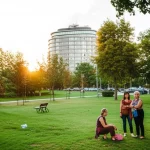 Fotografia grandangolare di un paesaggio urbano con aree verdi integrate, simboleggiante l'equilibrio tra ambiente costruito e naturale. Persone anziane che camminano serenamente nel parco in primo piano. Obiettivo grandangolare 24mm, luce del tardo pomeriggio, messa a fuoco nitida su tutto il piano, colori naturali.