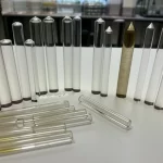Immagine fotorealistica di diversi campioni di vetro borato di zinco drogato con antimonio, di varie forme (dischi e bastoncini) e con diverse concentrazioni di antimonio che ne alterano leggermente il colore da trasparente a giallastro, disposti su un tavolo da laboratorio pulito. Lente macro 90mm, illuminazione da studio controllata per esaltare la trasparenza e le sfumature, alta definizione dei dettagli superficiali dei vetri, profondità di campo per mettere a fuoco alcuni campioni in primo piano.