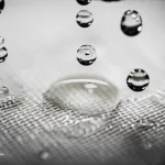 A close-up, macro lens shot at 60mm, showing water droplets passing through a fine filter membrane, highlighting the high detail and precise focusing on the filtration process.