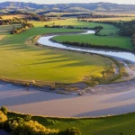 Veduta aerea di un fiume serpeggiante attraverso un paesaggio agricolo misto e aree boschive in Nuova Zelanda, l'acqua appare leggermente torbida suggerendo trasporto di sedimenti durante un evento di piena moderata. Fotografia paesaggistica, obiettivo grandangolare 24mm, luce naturale del tardo pomeriggio, messa a fuoco nitida su tutto il paesaggio, colori vividi.