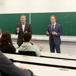 Aula universitaria moderna e luminosa con studenti divisi tra presenti fisicamente che interagiscono con un docente carismatico vicino a una lavagna interattiva e altri studenti collegati su grandi schermi murali, focus sullo schermo che mostra un modello ingegneristico 3D complesso, obiettivo 35mm, profondità di campo media per includere sia il docente che gli studenti, luce naturale brillante.
