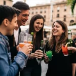 Gruppo di giovani amici italiani (uomini e donne, 20-25 anni) in un aperitivo all'aperto in una piazza italiana, alcuni uomini ridono fragorosamente con birre in mano, una donna nel gruppo sembra leggermente a disagio o giudicata mentre sorseggia un cocktail colorato, luce naturale del tardo pomeriggio che crea lunghe ombre, obiettivo 35mm, profondità di campo media per mostrare l'interazione e l'ambiente.