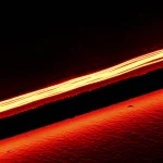 Immagine macro fotorealistica, obiettivo 90mm, alta definizione, illuminazione drammatica controllata, che mostra un singolo nanotubo di carbonio sospeso e incandescente che emette luce nel vicino infrarosso sopra una fessura microscopica in un substrato di silicio, enfatizzando l'emissione di luce non termica ad alta temperatura.