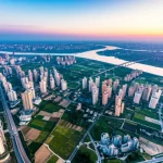 Veduta aerea mozzafiato della Cintura Economica del Fiume Azzurro, Cina, ripresa con un drone. Obiettivo grandangolare 10mm, lunga esposizione per evidenziare il corso sinuoso del fiume e le nuvole eteree, mettendo in risalto la complessa interazione tra aree urbanizzate, terreni agricoli e zone naturali. Luce dorata dell'alba che illumina il paesaggio, creando un forte contrasto e profondità.