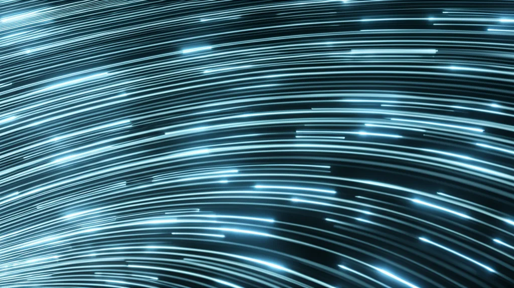 Visualizzazione artistica di un flusso di dati digitali astratti, con colori freddi come blu e argento, che si trasformano dinamicamente in una rappresentazione tridimensionale stilizzata della superficie di Fermi di un superconduttore. Particelle luminose, simboleggianti gli elettroni, si muovono senza resistenza apparente lungo questa superficie. L'immagine è catturata con un obiettivo grandangolare da 18mm, utilizzando una lunga esposizione per creare scie luminose eteree e un senso di movimento fluido, evocando la natura quantistica e l'efficienza della superconduttività.