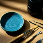 Un frammento di ceramica Jun della dinastia Yuan, con la sua caratteristica smaltatura blu-verdastra opalescente, appoggiato su un tavolo da archeologo. Accanto, strumenti di scavo come pennelli e piccole spatole. Macro lens, 80mm, high detail, precise focusing, controlled lighting, luce soffusa da una finestra laterale che crea ombre morbide.