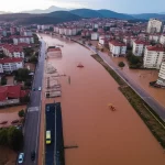 Immagine aerea drammatica di una città turca, Sinop o Kastamonu, parzialmente sommersa dall'acqua marrone e detritica di un'alluvione devastante. Si vedono tetti di case, strade trasformate in fiumi e squadre di soccorso che operano con gommoni. Wide-angle lens, 10-24mm, per catturare l'estensione del disastro, long exposure times per l'acqua, sharp focus sui dettagli della distruzione e dei soccorsi, luce del tardo pomeriggio per accentuare le ombre e il dramma della scena.