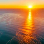 Panoramica mozzafiato di una costa vista dall'alto al tramonto, con le correnti oceaniche visibili come pattern nell'acqua illuminata dal sole, obiettivo grandangolare 20mm, lunga esposizione per nuvole e acqua setose, colori caldi.