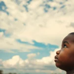 Immagine suggestiva che rappresenta il legame tra fattori meteorologici e la salute dei bambini in Etiopia: un bambino piccolo guarda verso un cielo mutevole, con un filtro duotone seppia e blu per un effetto emotivo. Obiettivo da ritratto 50mm, profondità di campo media.