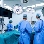 Fotografia ad angolo ampio di una moderna sala operatoria durante un intervento di ernia inguinale TEP. Il team chirurgico è concentrato sugli schermi laparoscopici. Luce brillante e sterile, equipaggiamento high-tech visibile. Obiettivo grandangolare 24mm, messa a fuoco nitida.