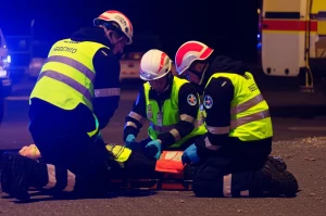 Fotografia realistica in stile reportage di un team di soccorso tedesco (medici e paramedici) che applica un algoritmo di triage su un paziente simulato durante un'esercitazione notturna di maxi-emergenza. Teleobiettivo zoom 100-400mm, fast shutter speed per catturare l'azione, illuminazione drammatica dalle luci di emergenza, focus nitido sui volti concentrati e sulle schede di triage.